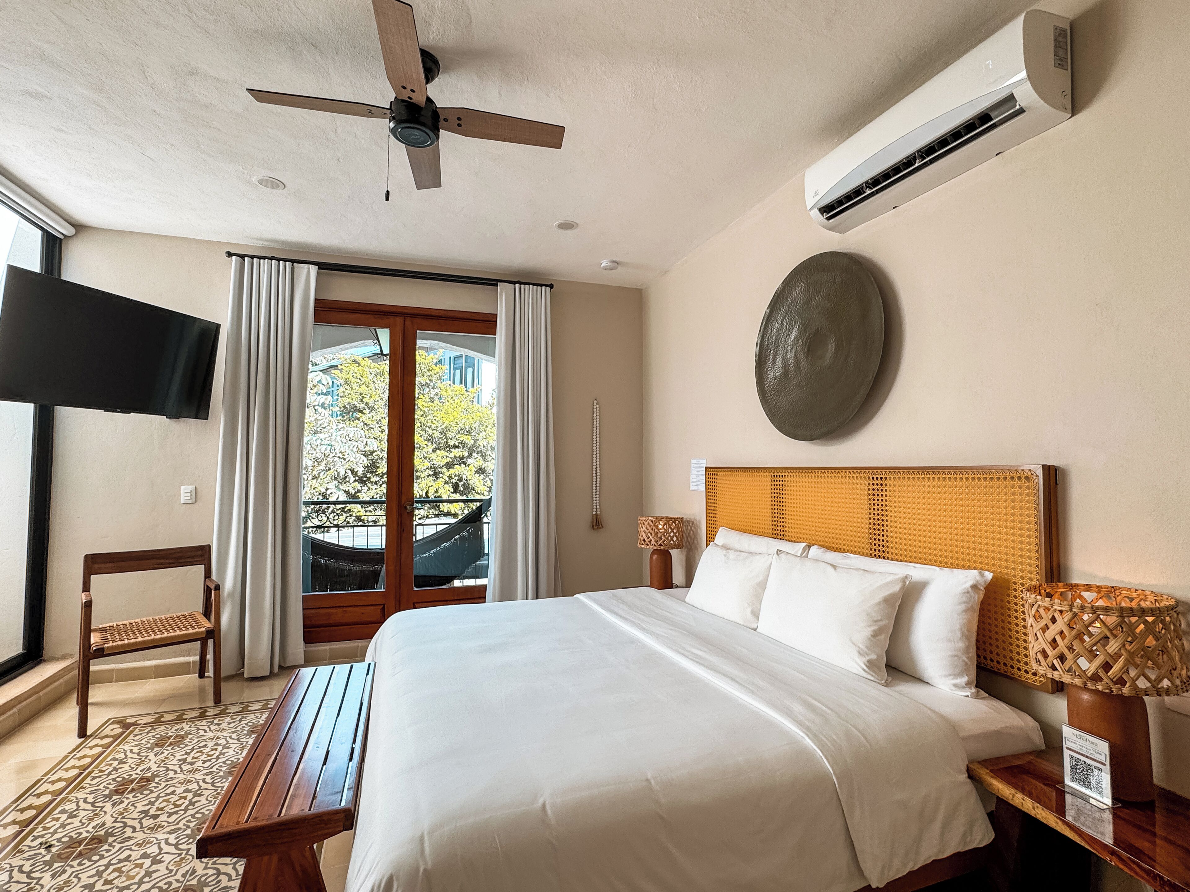Habitación de hotel con cama, ventilador de techo, aire acondicionado, televisión, ventana con cortinas y vista al exterior, además de un adorno en la pared.