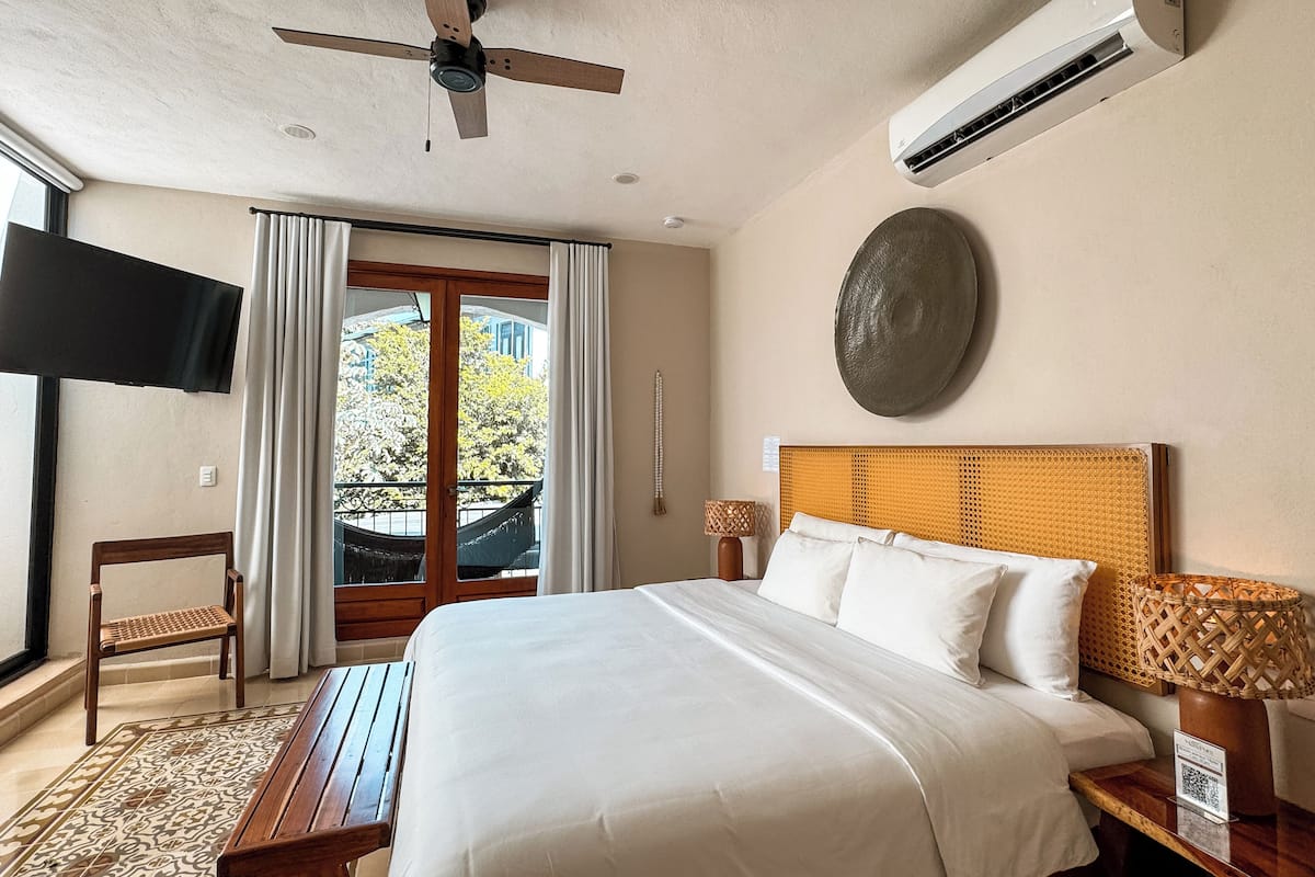 A hotel room with a bed, a ceiling fan, an air conditioning unit, a TV, a window with curtains, a view of the outside, and a decorative wall piece.