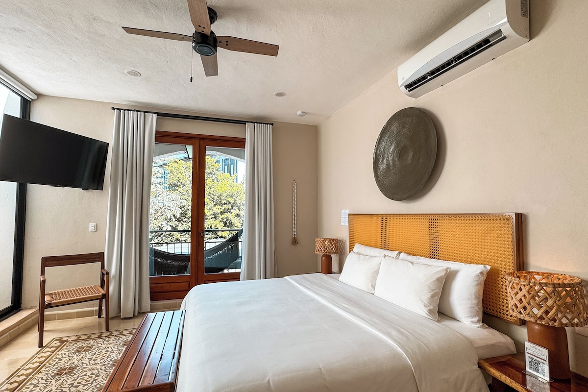 Habitación de hotel con cama, ventilador de techo, aire acondicionado, televisión, ventana con cortinas y vista al exterior, además de un adorno en la pared.
