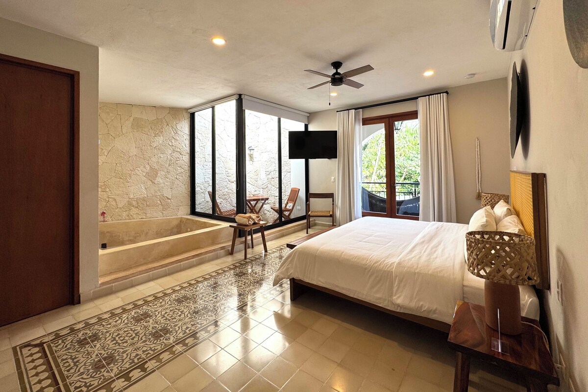 A spacious bedroom with a large bed, a sitting area with chairs, a bathtub, and a balcony with curtains.