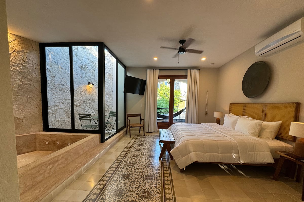 A hotel room with a large bed, a bathtub, a TV, and a balcony with a view of greenery.