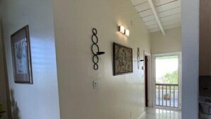 A hallway with white walls, tiled flooring, and a ceiling with exposed beams. There are three wall-mounted light fixtures and two framed artworks. - Angie's Sunset View (St Thomas)