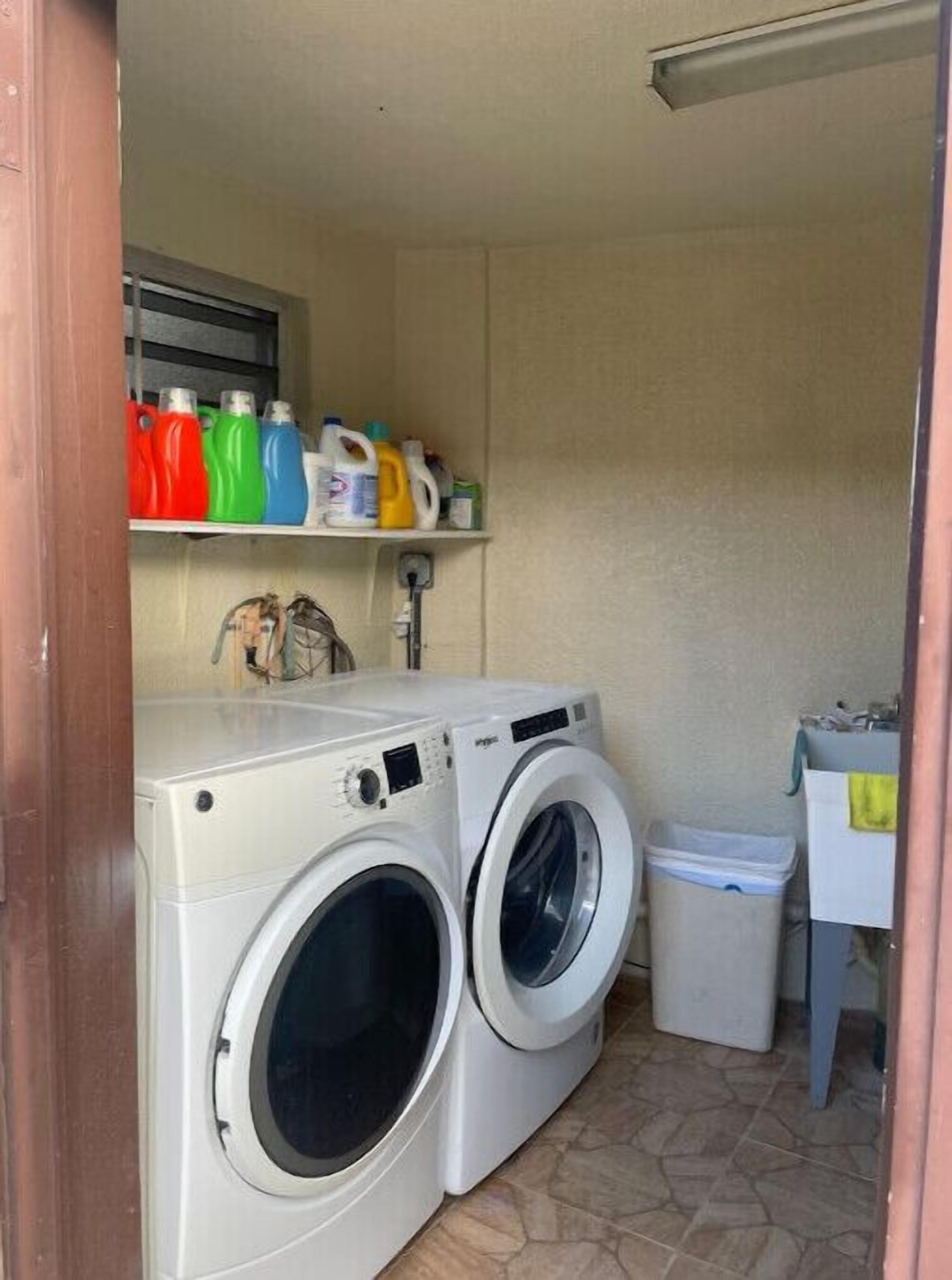 A laundry room with two front-loading washing machines, a shelf with various laundry detergents, and a trash bin.