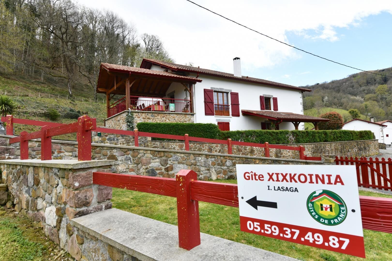 A white building with red accents, a stone wall, and a red fence. A sign indicates the location as Gîte XIXKOINIA, I. LASAGA, with a phone number.