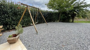 Een grindterras met een houten schommel, omringd door dichte heggen en een grote boom.