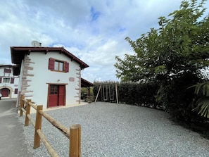 Um edifício rústico de pedra com porta vermelha e persianas, um caminho de acesso de cascalho e uma cerca de madeira.
