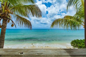 A wooden deck overlooking a beach with clear turquoise water, palm trees, and a distant boat.