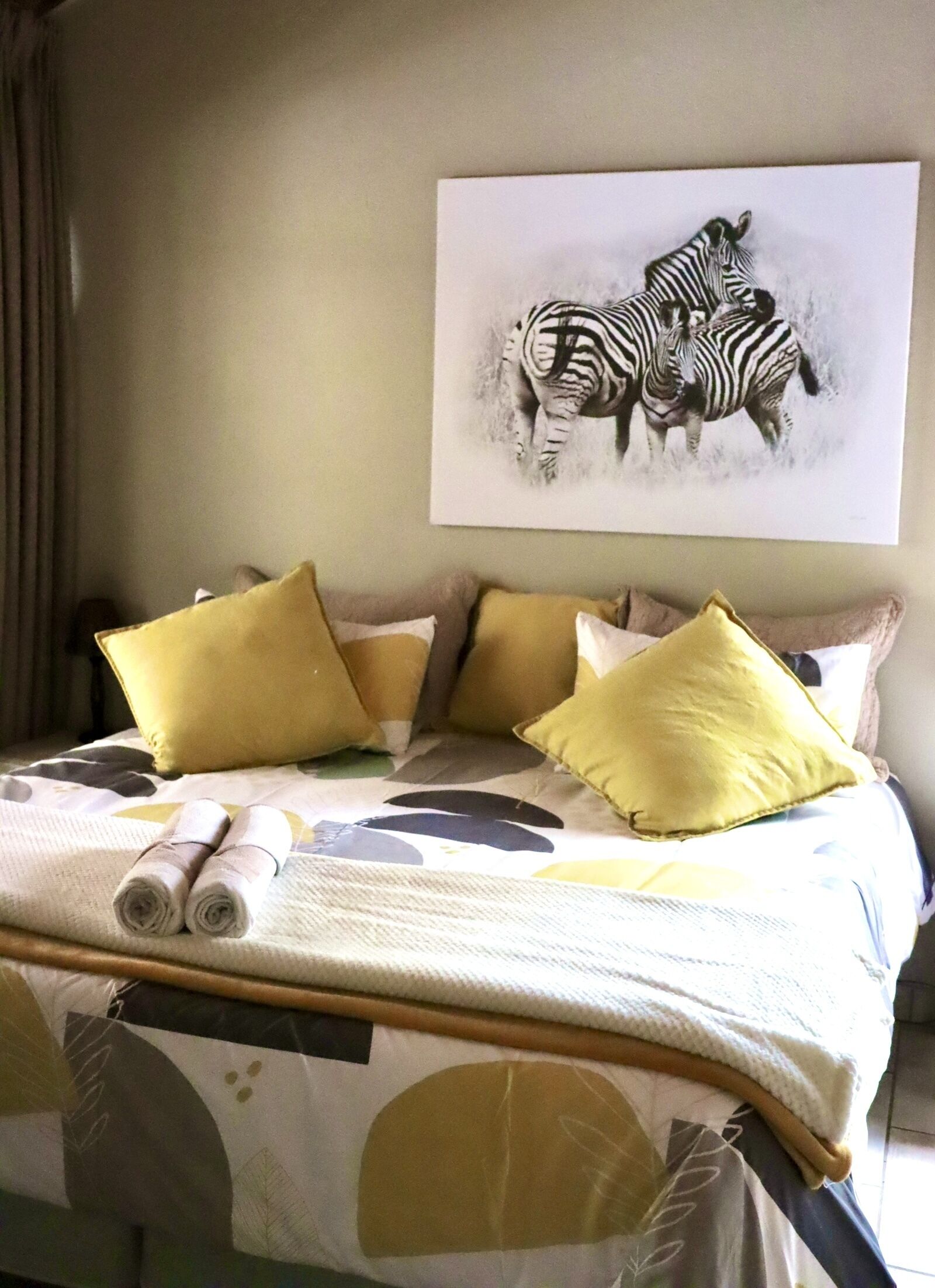 A neatly made bed with a patterned bedspread, two rolled towels, and multiple pillows in a room with a plain wall and a framed zebra artwork.