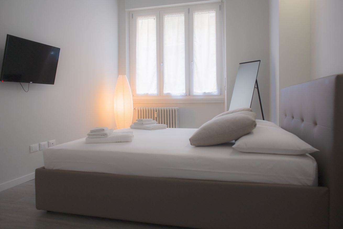 Een slaapkamer met een bed, een bedlamp, een flatscreen televisie, een raam met zonwering en een radiator.