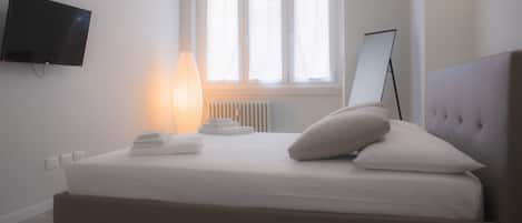 A bedroom with a bed, a bedside lamp, a flat-screen TV, a window with blinds, and a radiator.
