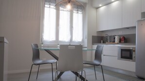 Une cuisine moderne avec une table à manger en verre, deux chaises, un luminaire suspendu, une fenêtre avec des rideaux légers et des appareils électroménagers intégrés.