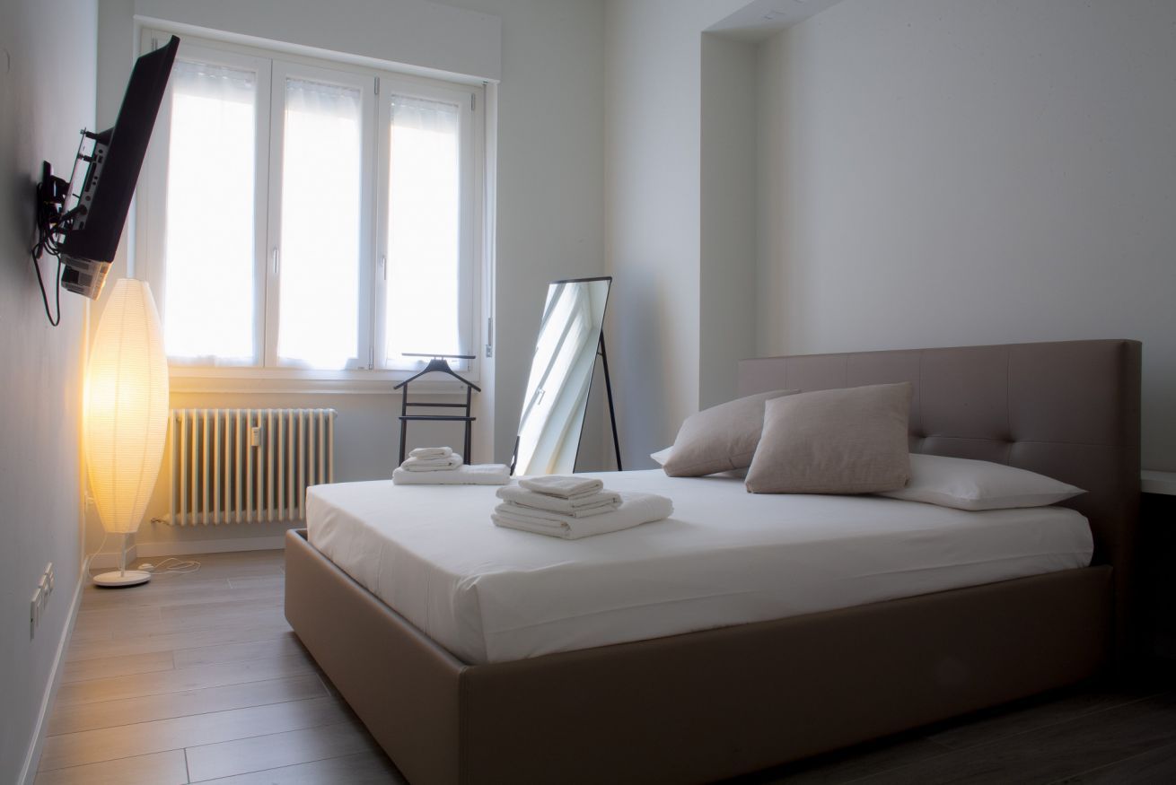 A bedroom with a bed, a radiator, a standing mirror, a TV mounted on the wall, and a window with blinds.