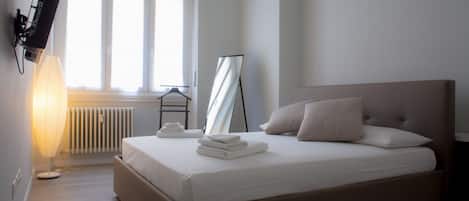 A bedroom with a bed, a radiator, a standing mirror, a TV mounted on the wall, and a window with blinds.