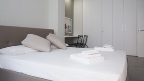 Une chambre à coucher moderne avec un grand lit, une tête de lit, une table de chevet, une chaise, une armoire et une petite œuvre d’art accrochée au mur.