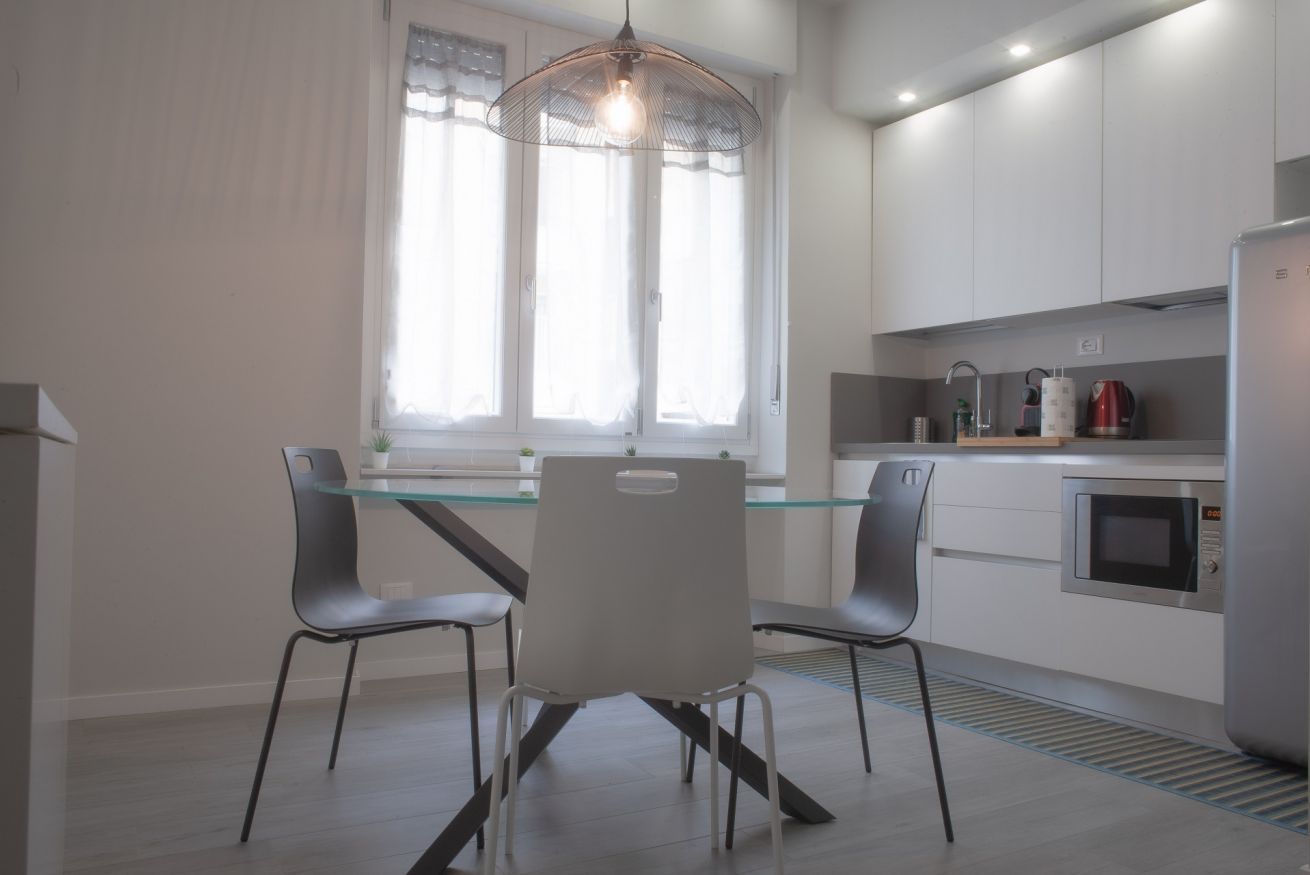 Een moderne keuken met een glazen eettafel, twee stoelen, een hanglamp, een raam met dunne gordijnen en ingebouwde apparatuur.