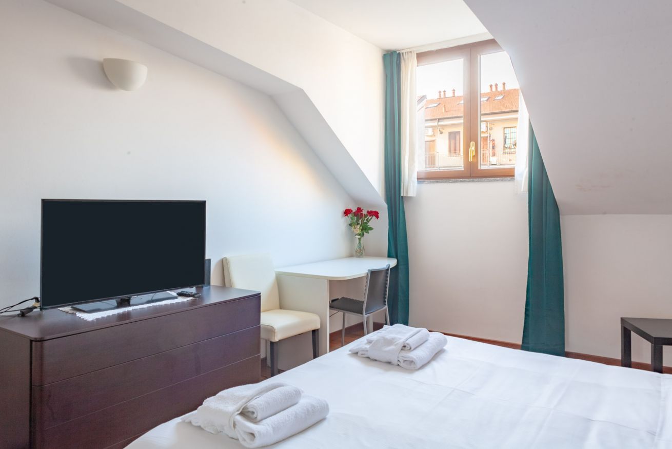 A hotel room with a bed, a TV, a desk, and a window with curtains.