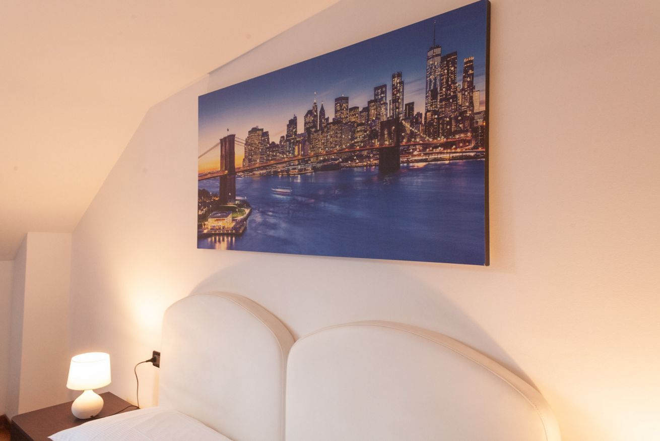 Ein Hotelzimmer mit Stadtbildgemälde, einem Bett und einem Nachttisch mit Lampe.