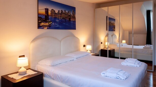 A hotel room with a large bed, a nightstand with a lamp, a mirror, and a cityscape painting on the wall. - ASIAGO, 41 · Quartiere Gorla, verde in città (Milano)