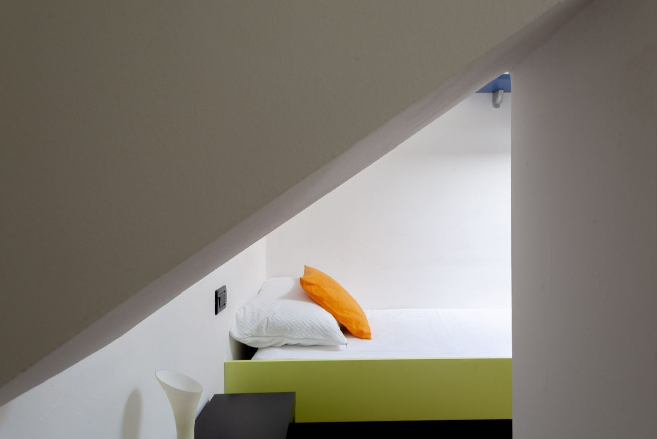 Una cama individual superpuesta con un cojín blanco y naranja, un florero blanco y un objeto negro en el suelo.