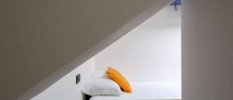 Una cama individual superpuesta con un cojín blanco y naranja, un florero blanco y un objeto negro en el suelo.