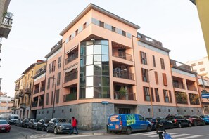Een meerlaags gebouw met balkons, een blauwe bestelwagen geparkeerd ervoor en een persoon die over het trottoir loopt.