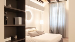 A bedroom with a bed, a bedside table, a lamp, a shelf with books and a decorative item, and a window with curtains. - VIA SIMONE MAYR, 2 · Bergamo città Alta - appartamento di charme (Bergamo)