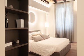 A bedroom with a bed, a bedside table, a lamp, a shelf with books and a decorative item, and a window with curtains. - VIA SIMONE MAYR, 2 · Bergamo città Alta - appartamento di charme (Bergamo)