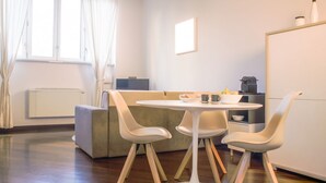 A modern living room with a round white table, four white chairs, a grey sofa, a window with curtains, a radiator, and a cabinet with kitchen appliances.