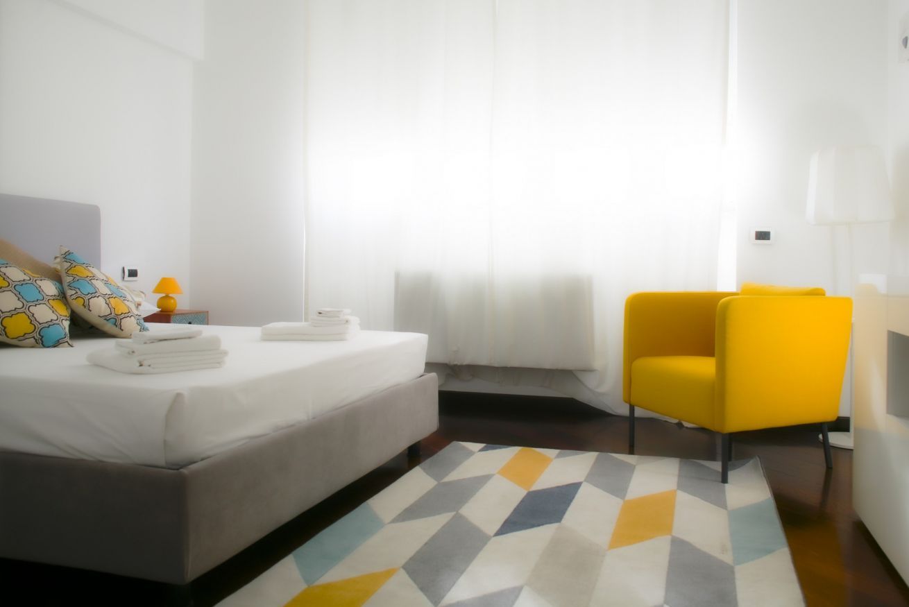 A hotel room with a bed, a yellow armchair, a bedside table with a lamp, and a geometric-patterned rug.