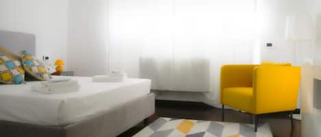 Habitación de hotel con cama, un sillón amarillo, mesita de noche con lámpara y alfombra de estampado geométrico.