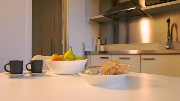 A kitchen with a white countertop, a bowl of fruit, a cup, and a plate of food.