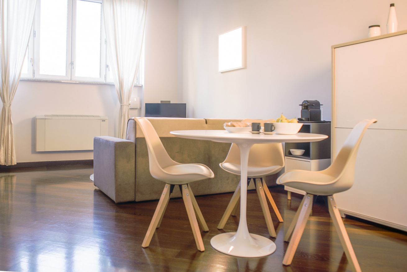 Een moderne woonkamer met een ronde witte tafel, vier witte stoelen, een grijze bank, een raam met gordijnen, een radiator en een kast met keukenapparatuur.