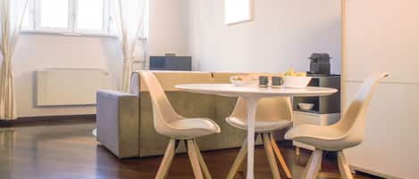 A modern living room with a round white table, four white chairs, a grey sofa, a window with curtains, a radiator, and a cabinet with kitchen appliances.