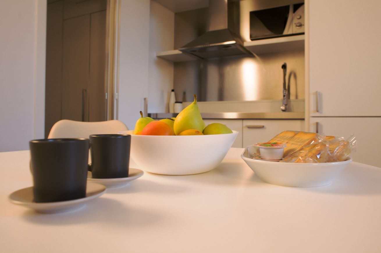 A kitchen with a white countertop, a bowl of fruit, two black cups, and a plate with bread.