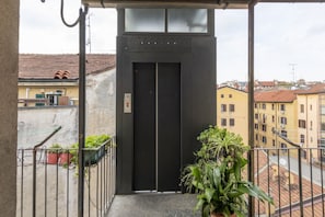 An elevator entrance with a metal door, a potted plant to the right, and a view of buildings in the background.