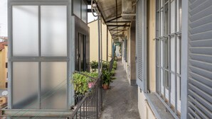 A narrow corridor with a metal railing, potted plants, and a series of windows on one side. - Buenos Aires 49 – Lungo la via dello shopping (Milano)