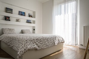 Un dormitorio con cama, pared blanca con estantes y ventana con cortinas traslúcidas.