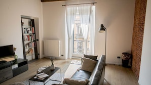 Un salon comprenant un canapé, une table basse, une bibliothèque, un radiateur et une fenêtre avec des rideaux.
