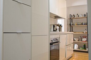 Una cocina con muebles blancos, horno empotrado y estante con frascos y utensilios de cocina.