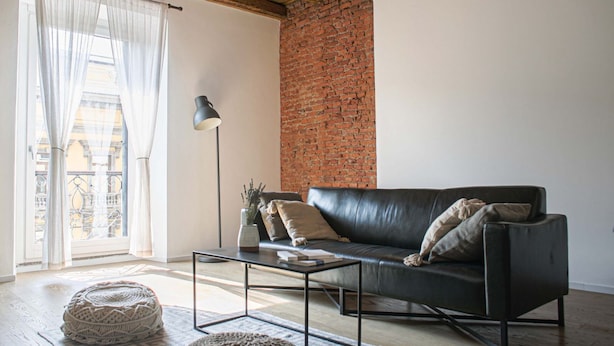 Un salon avec un canapé en cuir noir, une table basse, une lampe et une vue sur l’extérieur à travers une fenêtre ornée de rideaux légers.