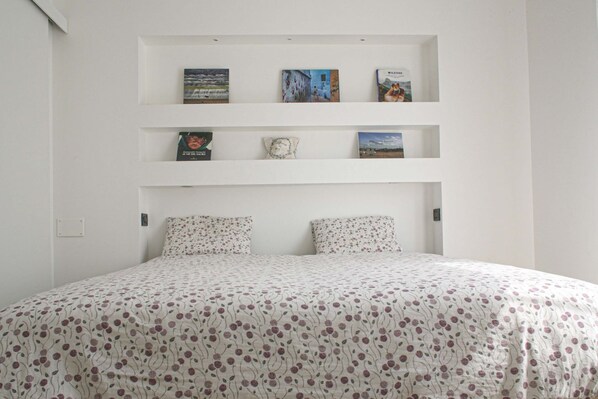 A bedroom with a bed, two pillows, a white wall, and three shelves with books and a clock.