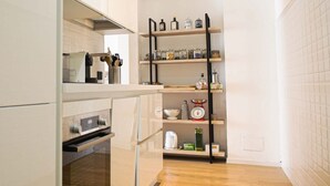 Une cuisine compacte équipée d’un four encastré, d’une machine à café et d’une étagère où sont rangés des bocaux et des ustensiles de cuisine.