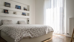 Une chambre à coucher avec un lit, un mur blanc orné d’étagères et une fenêtre avec des rideaux légers.