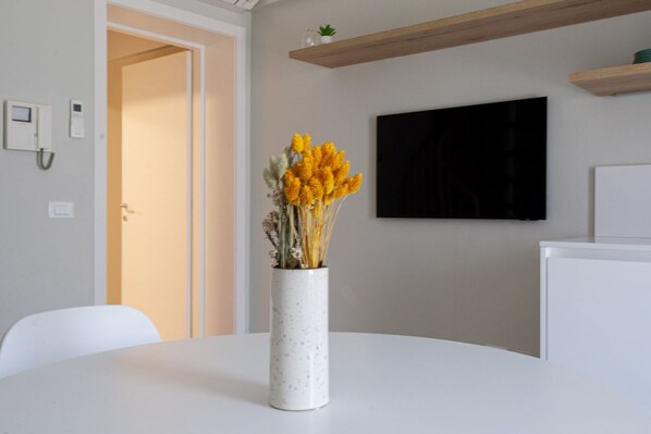A white table with a vase of yellow flowers, a white chair, a door, a mounted TV, and a wooden shelf. - CASATI, 2 · Glamour in città tra shopping e vita notturna (Milano)