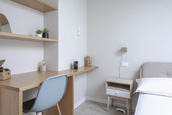 A bedroom with a bed, a desk, a chair, a nightstand, and a wall-mounted shelf with decorative items.