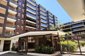 A modern multi-story residential building with balconies, a covered entrance, and a gated parking area.