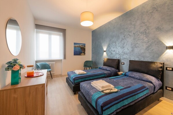A hotel room with two beds, a wooden desk, a mirror, a window with a view, and a wall-mounted light fixture.