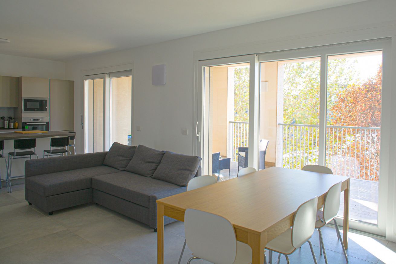 Un espacio habitable moderno con un sofá modular, una mesa de comedor y sillas. Hay una zona de cocina con un microondas y una mesa de comedor. Amplias ventanas ofrecen vistas al exterior.
