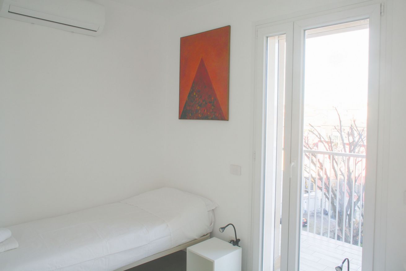 Une chambre avec un lit simple, un matelas blanc, une petite table de chevet, un tableau accroché au mur et une porte-fenêtre coulissante donnant sur un espace extérieur.