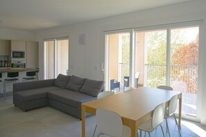 A modern living space with a sectional sofa, a dining table, and chairs. There is a kitchen area with a microwave and a dining table. Large windows provide a view of the outdoors.